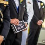A group of men in formal military and business attire holding a 'Top Secret' briefcase indoors.